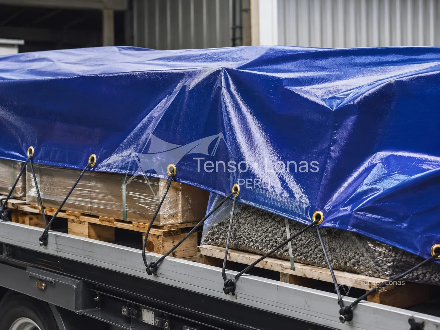 Manta de tarflex azul cubriendo carga paletizada sobre la plataforma de un camión, sujeta con tensores por los ojales