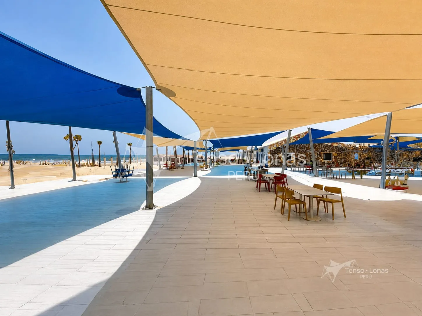 Toldos tipo vela beige y azules sobre la terraza de un comercio frente al mar